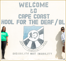 Sierra and a staff member of the Cape Coast School for the Deaf and Blind stand in front of the painted mural showing their logo, a coat of arms that is diagonally cut into a blue sectional and striped black and white sectional with a drawing of a walking man with a cane and an ear in the middle. The bottom reads 'Disability not inability'